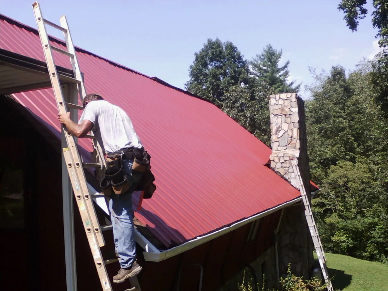 Roofer on a ladder beside a red metal roof and stone chimney — Soffit & Fascia Repair by Roofing Brothers in Streetsboro